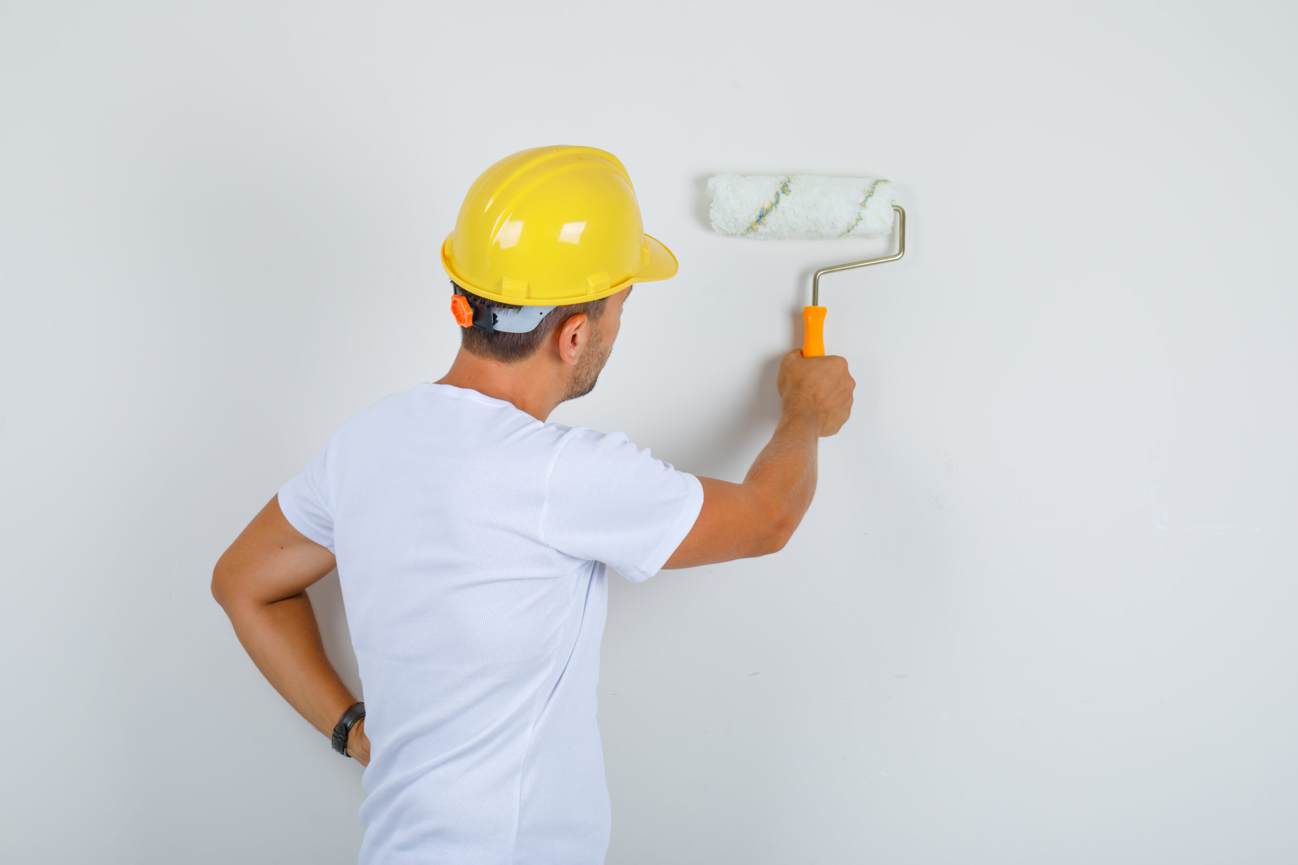 builder man painting wall with roller in white t shirt, helmet and looking busy , back view.